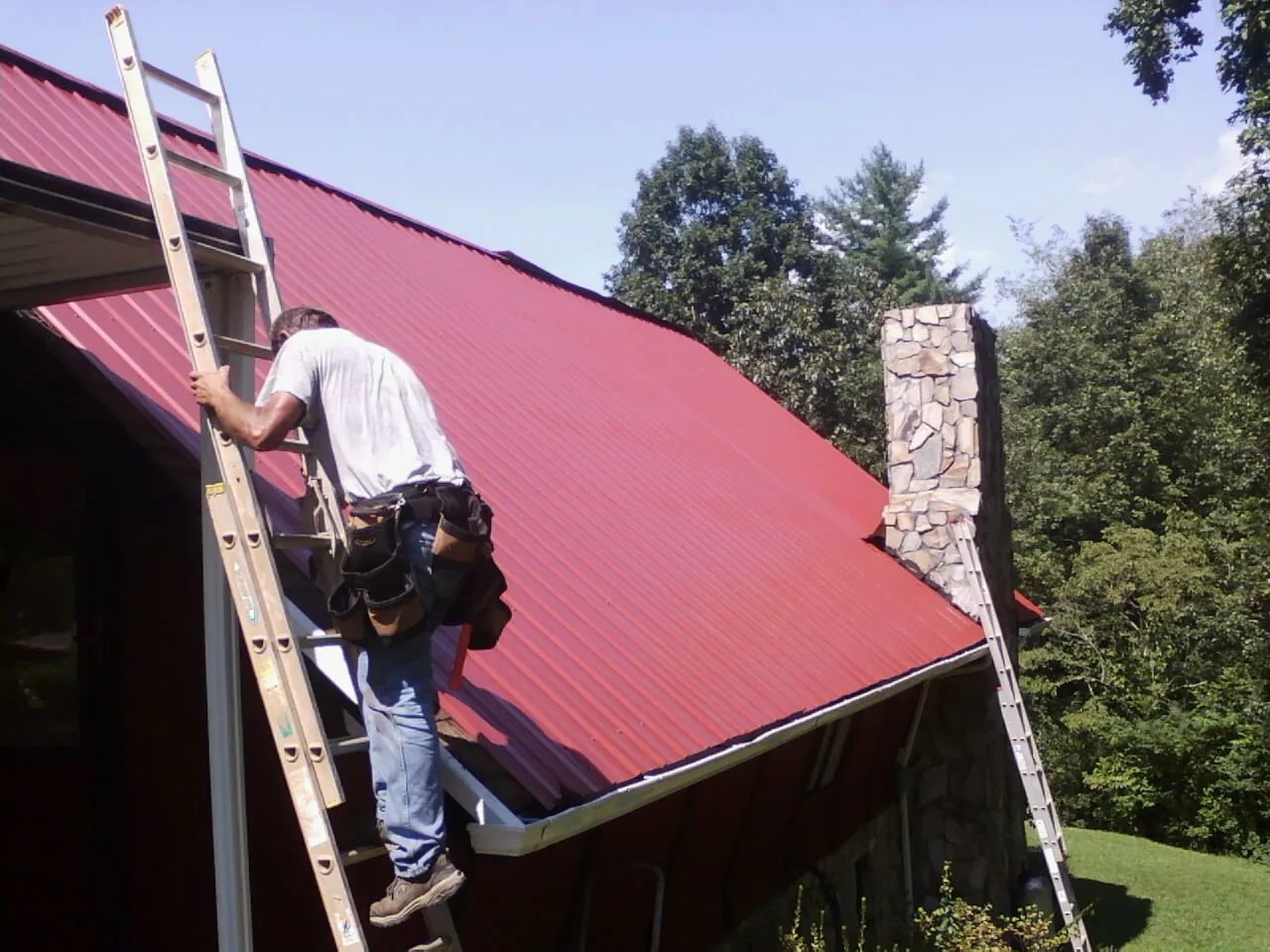 Roofer on a ladder beside a red metal roof and stone chimney — Soffit & Fascia Repair by Roofing Brothers in Thousand Oaks